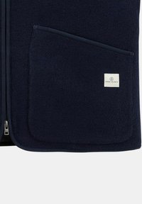 Gilet en polaire bleu marine avec une fermeture éclair à l'avant, grande poche et petite étiquette de logo. Texture douce et design simple et fonctionnel.