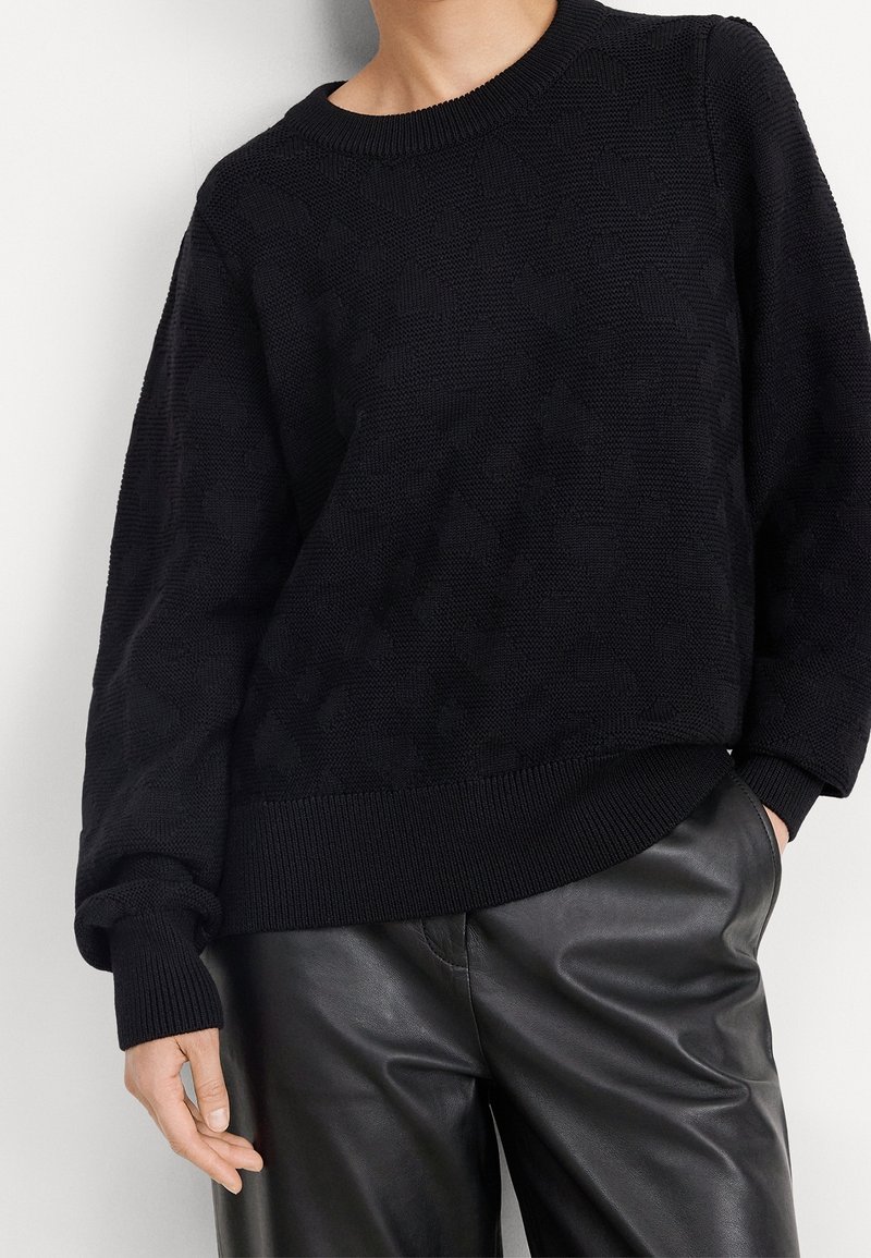 Suéter negro de punto con un patrón texturizado, escote redondeado y puños de canalé. Combinado con pantalones de cuero negros.