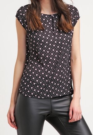 Frau trägt ein schwarzes Kurzarm-Top mit pink-weißem Rautenmuster und schwarze Lederhose, gezeigt von den Schultern bis zu den Oberschenkeln.