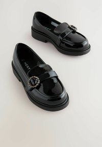 Loafers negri din patent cu talpă plată, având o curea decorativă și un accent de piatră circulară în față. Finisaj neted și strălucitor.