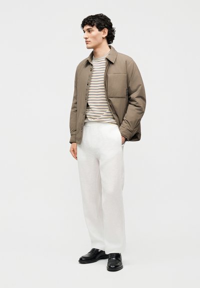 Άνδρας που φοράει καφέ πουκάμισο με κουμπιά, ριγέ μπεζ και άσπρο πουκάμισο, λευκό παντελόνι και μαύρα παπούτσια, στέκεται με τα χέρια στις τσέπες.