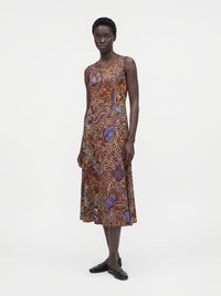 Femme debout portant une robe midi marron sans manches avec des motifs floraux et abstraits bleus et orange, associée à des chaussures plates noires.