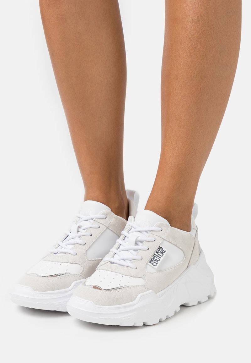 Fötter som bär vita chunky sneakers med mocka- och läderpaneler, märkta "Versace Jeans Couture" på sidan.