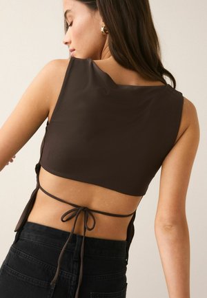 Femme aux longs cheveux foncés portant un top court marron sans manches noué dans le dos, associé à un jean noir taille haute.