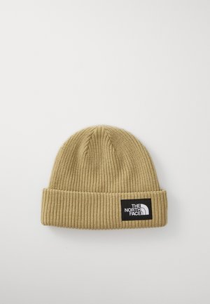Tan knitted beanie with a ribbed texture, featuring a black rectangular logo patch on the front. The design is simple and functional.