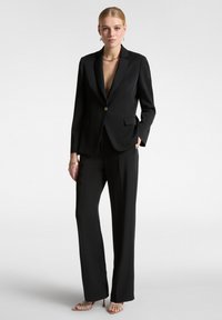 Completo nero su misura con giacca fitted e pantaloni a gamba larga, caratterizzati da un unico bottone dorato e tasche frontali. Tessuto liscio e leggero.