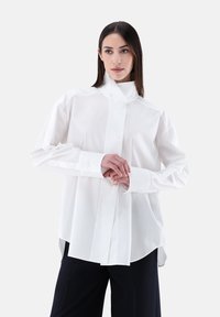 Camicia bianca a maniche lunghe con colletto alto, realizzata in un tessuto liscio, con chiusura frontale con bottoni e dettagli sul patta, vestibilità ampia e spacchi laterali.