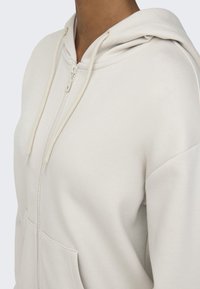 Cream-colored zip-up hoodie with a textured fabric, front pockets, and drawstring hood, featuring a seamless design and minimal stitching.