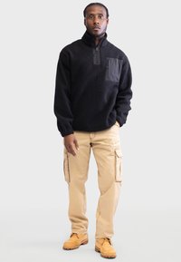 Pulover negru din fleece cu fermoar pe un sfert, având o suprafață texturată și un buzunar pe piept din material negru. Asortat cu pantaloni cargo bej și cizme.