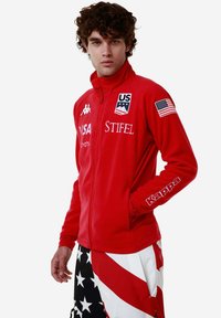 Giacca rossa con zip e bandiera degli Stati Uniti con loghi del marchio. Tessuto morbido, colletto alto, design aderente. Presenta dettagli bianchi sulle maniche. Aspetto sportivo distintivo.