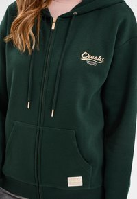 Grüner Hoodie mit einer Vordertasche, goldfarbenem Reißverschluss und gesticktem "Creeks"-Logo. Hergestellt aus weichem Stoff mit glatter Textur.