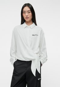 Camisa blanca de manga larga, tipo envoltura, con cuello y detalle de cintura atada. Presenta un logo bordado en negro. Tejido suave con un corte relajado.