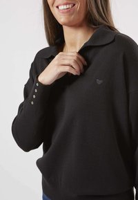 Pull-over noir avec col, arborant un petit logo en forme de cœur et des détails de boutons sur les manches. Texture douce et coupe décontractée.