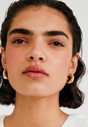 Kuldsest rõngaskujulised kõrvarõngad tekstureeritud pinnaga, kantud inimesel, kellel on lühikesed lokkis juuksed, selge nahk ja neutraalne särk, neutraalne taust.