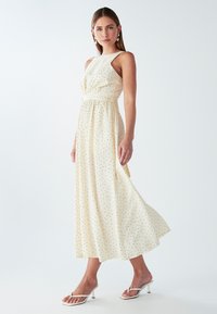 Žena u bezrukavoj krem haljini midi dužine s crnim točkicama i bijelim sandalama na петu, stoji uz jednostavnu pozadinu.