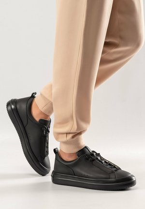 Schwarze Leder-Sneaker mit strukturiertem Finish, Schnürdesign und kontrastierender Gummisohle, kombiniert mit beigen, schmal geschnittenen Hosen.