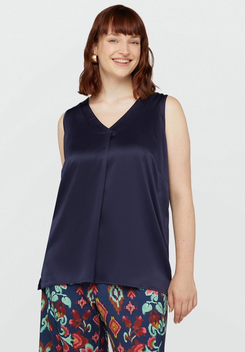 Top senza maniche in satin blu navy con scollo a V e leggera drappeggiatura, caratterizzato da una piega frontale. Abbinato a pantaloni colorati e decorati.