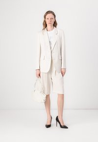 Blazer ajusté de couleur claire sur un t-shirt graphique, associé à un short crème et des talons noirs. Le mannequin tient un petit sac à main blanc.