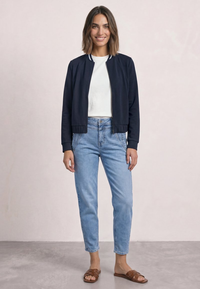 Woman standing smiling wearing navy jacket, white shirt, light blue jeans, and brown sandals against plain light background.
