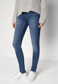Jeans ajustados en denim azul medio, con una textura suave, desgaste sutil y un diseño ceñido al cuerpo. Combinados con zapatillas blancas.