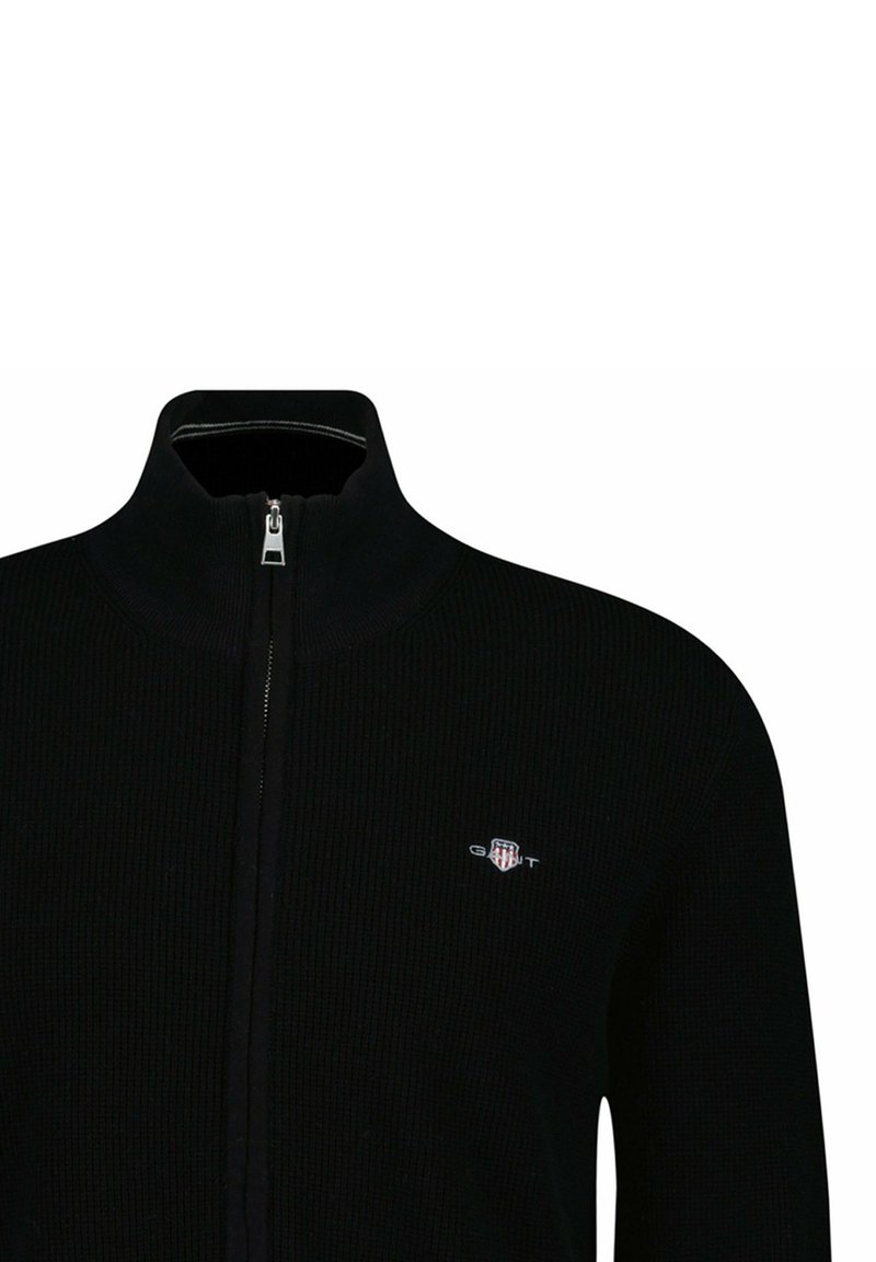 Suéter negro con cremallera hecho de tela acanalada, con cuello alto y un pequeño logo bordado en el lado izquierdo.