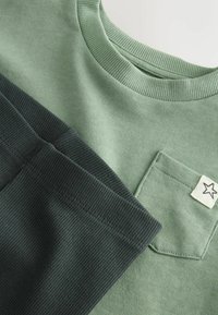 Grünes und dunkelgrünes Baumwoll-T-Shirt mit einem runden, gerippten Ausschnitt und Tasche. Verfügt über ein kleines Stern-Patch auf der Tasche.