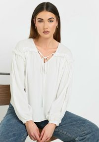 Hvid bluse med båndhale-detalje, bløde ærmer og flæsedetaljer ved skuldrene. Blødt stof og afslappet pasform, parret med denim.