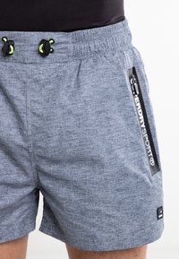 Šedé sportovní šortky s texturovaným vzorem, elastickým pasem a černou šňůrkou na stažení. Obsahují postranní logovou nášivku a průhledný detail kapsy.