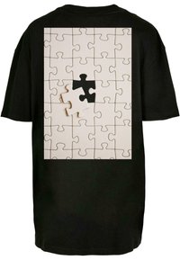 Sort oversized t-shirt med et stort trykt puslespildesign i hvid og sort, der viser et manglende brik på ryggen.