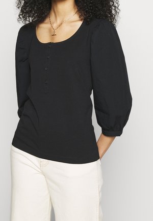 Kobieta w czarnej bluzce z guziczkami, rękawy 3/4 z bufkami, i kremowe spodnie, z kręconymi włosami i złotym naszyjnikiem.