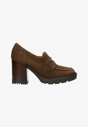 Mocassins à talons en suède brun avec un bout arrondi, dotés d'une sangle décorative, d'un talon bloc texturé et d'une semelle rugueuse pour une meilleure adhérence.