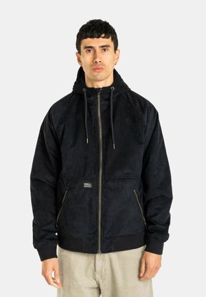 Mann mit kurzen dunklen Haaren, der eine schwarze Zip-up Kapuzenjacke und beige Hose trägt, steht vor einem schlichten hellen Hintergrund.