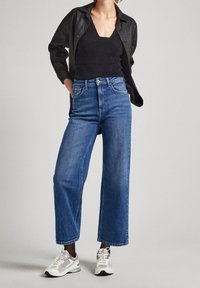 Korkeavyötäröiset siniset farkut, joissa on leveä lahje, yhdistetty mustan topin ja valkoisten-hopeisten lenkkareiden kanssa. Rakenteellinen kangas ja näkyvät sauman yksityiskohdat.