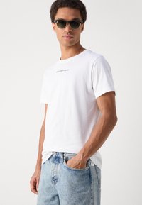 White cotton t-shirt with short sleeves, featuring a small black "Calvin Klein Jeans" logo on the chest, paired with light blue denim jeans.