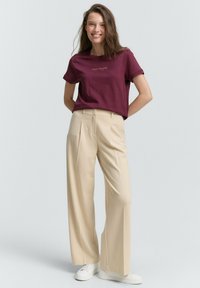 T-shirt à col rond bordeaux avec le texte "TOM TAILOR", associé à un pantalon large beige et des baskets blanches. Tissu lisse, coupe décontractée.