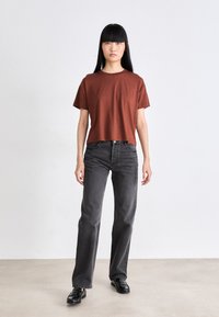 Roodkleurig t-shirt met korte mouwen gecombineerd met hoog opgesneden, rechte donkergrijze jeans. Model staat op een neutrale ondergrond.