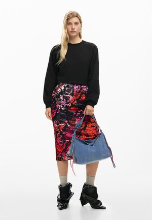 Vrouw in een zwarte trui en kleurrijke bloemenrok met een blauwe handtas, beige sokken en zwarte puntige laarzen, staand op een witte achtergrond.