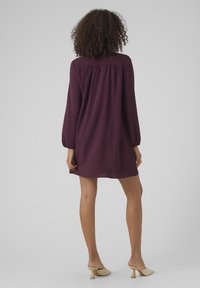 Bordeauxrotes Kleid mit langen Ärmeln, gerafftem Vorderteil, lockerer Passform, knielang, kombiniert mit hellen, hochhackigen Mules.