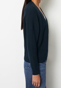 Vrouw die zijwaarts staat, gekleed in een donkerblauw gebreid vest over een witte top met blauwe spijkerbroek, tegen een effen witte muur.