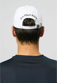 Baltas beisbolo kepurė su juodu siuvinėtu užrašu "U.S. POLO ASSN." nugaros dalyje. Reguliuojama juosta gale. Dėvima su tamsia marškinėliais.