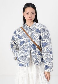 Quiltana jakna v beli barvi z modrimi paisley in cvetličnimi vzorci, z okroglim ovratnikom, sprednjim gumbom na zapiranje in stranskimi žepi.