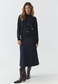 Marineblauwe gebreide trui met kleurrijke borduursels van kersen, sterren en bloemen, gecombineerd met een geplooide marineblauwe rok en bruine cowboylaarzen.