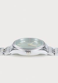 Montre en métal argenté avec une lunette texturée, un cadran vert et des aiguilles dorées. Elle présente un bracelet en acier inoxydable à maillons et une couronne.