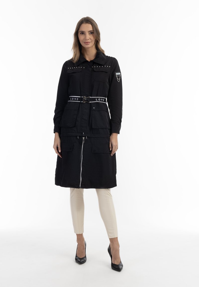 Giubbotto nero con vita abbottonata, con tasche a pressione, dettagli con borchie e zip frontale. Indossato sopra pantaloni beige e tacchi neri.