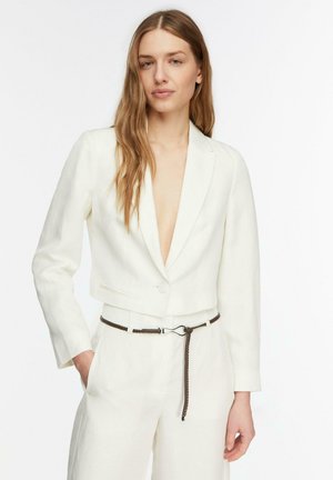 Kobieta o długich, jasnobrązowych włosach, ubrana w biały krótki blezer i spodnie, uzupełniona cienkim plecionym brązowym paskiem.