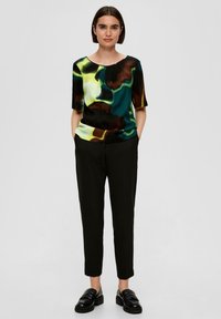 Chemisier à motifs floraux avec des manches courtes en vert, noir et jaune ; associé à un pantalon noir ajusté et des mocassins noirs.
