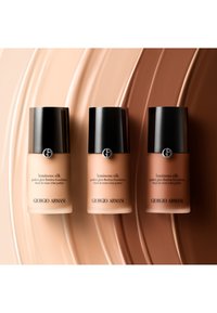 Tre flasker med Giorgio Armani Luminous Silk foundation i forskjellige nyanser; glassbeholdere med dype svarte lokk og elegante etiketttekster.