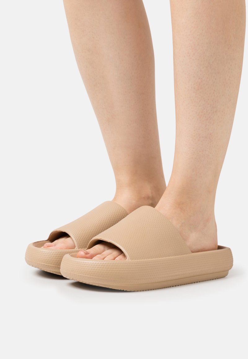 Beige Slipper-Sandalen mit strukturiertem Obermaterial, breitem Riemen und dicker Sohle, die die Zehen freilegen und eine glatte Gummi-Oberfläche haben.