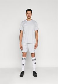 Γκρι αθλητική φανέλα και σορτς, κατασκευασμένα από ύφασμα που απομακρύνει την υγρασία, με λευκές λεπτομέρειες και τριστιβικό σήμα. Μαύρα ποδοσφαιρικά παπούτσια και ριγέ κάλτσες.