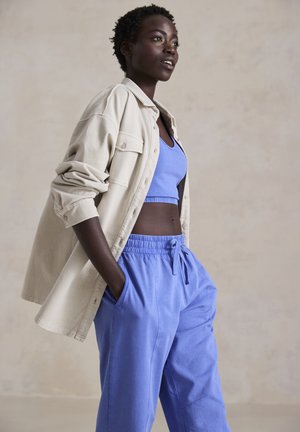 Lichtbeige shirtjack over een blauwe crop top en blauwe broek met koordjes. Glad stof, relaxte pasvorm en zijzakken.